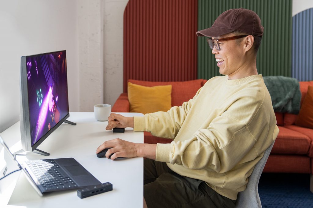 Un hombre con un suéter amarillo juega mientras usa los accesorios adaptativos de Microsoft. El hombre está sentado en una silla frente a un monitor que muestra un juego de carreras mientras usa el mouse adaptativo con la mano izquierda y el botón adaptativo con la mano derecha.