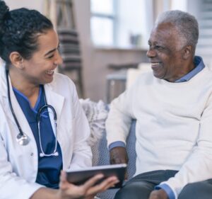 Uma mulher médica e negra à esquerda está vestindo um jaleco branco e segura um dispositivo eletrônico ao conversar sentada com um homem negro à direita que veste uma blusa branca.