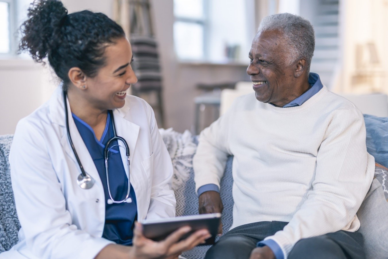 Uma mulher médica e negra à esquerda está vestindo um jaleco branco e segura um dispositivo eletrônico ao conversar sentada com um homem negro à direita que veste uma blusa branca.
