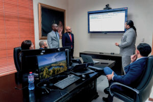 Seis personas reunidas en una sala de juntas observan una pantalla.