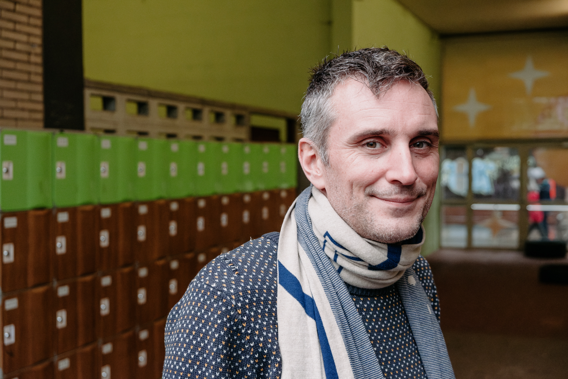 Un hombre con sueter y bufanda frente a unos casilleros en el pasillo de una escuela