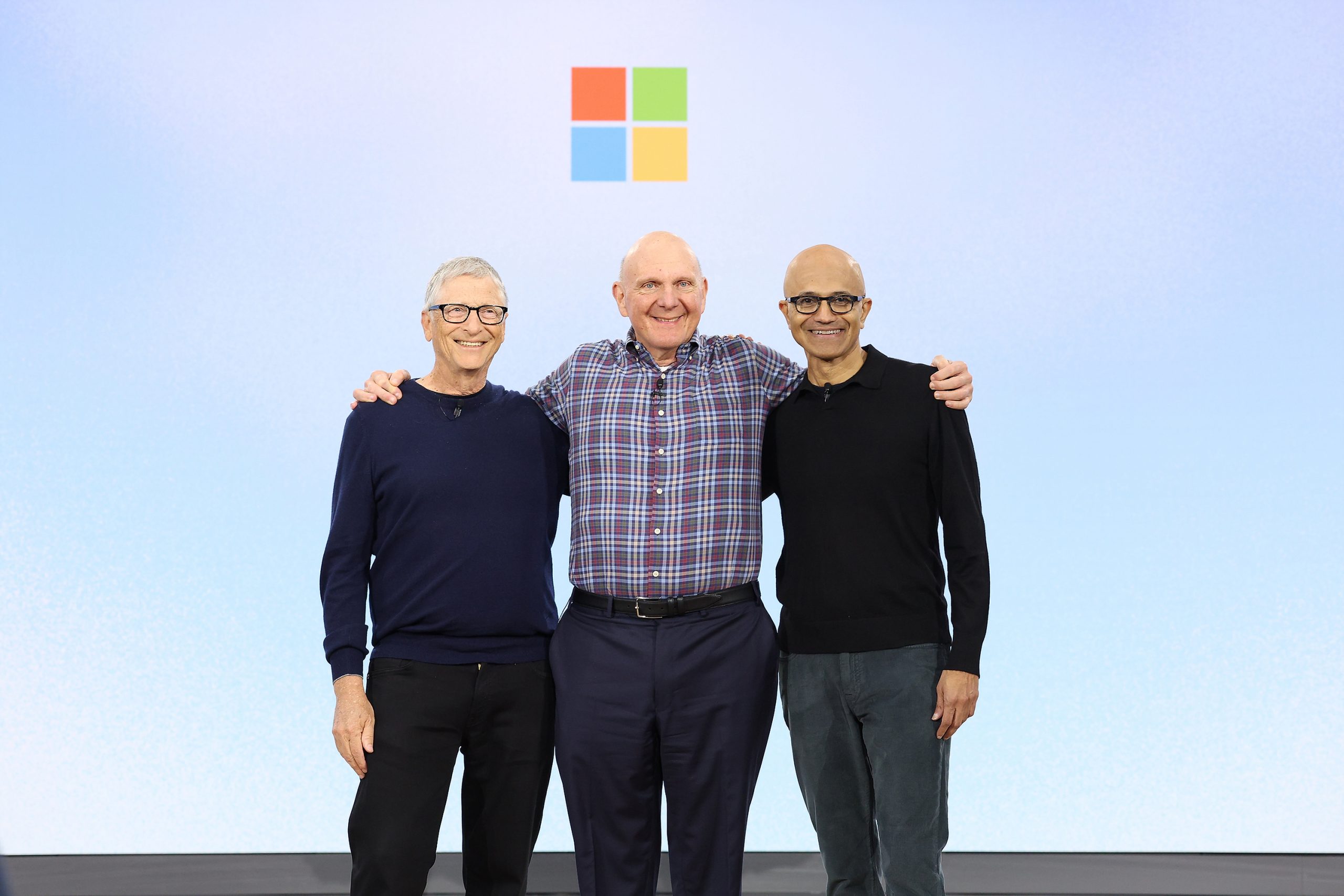 Tres hombres con los brazos alrededor de los hombros de cada uno están de pie en el escenario frente al colorido logo cuadrado de Microsoft.
