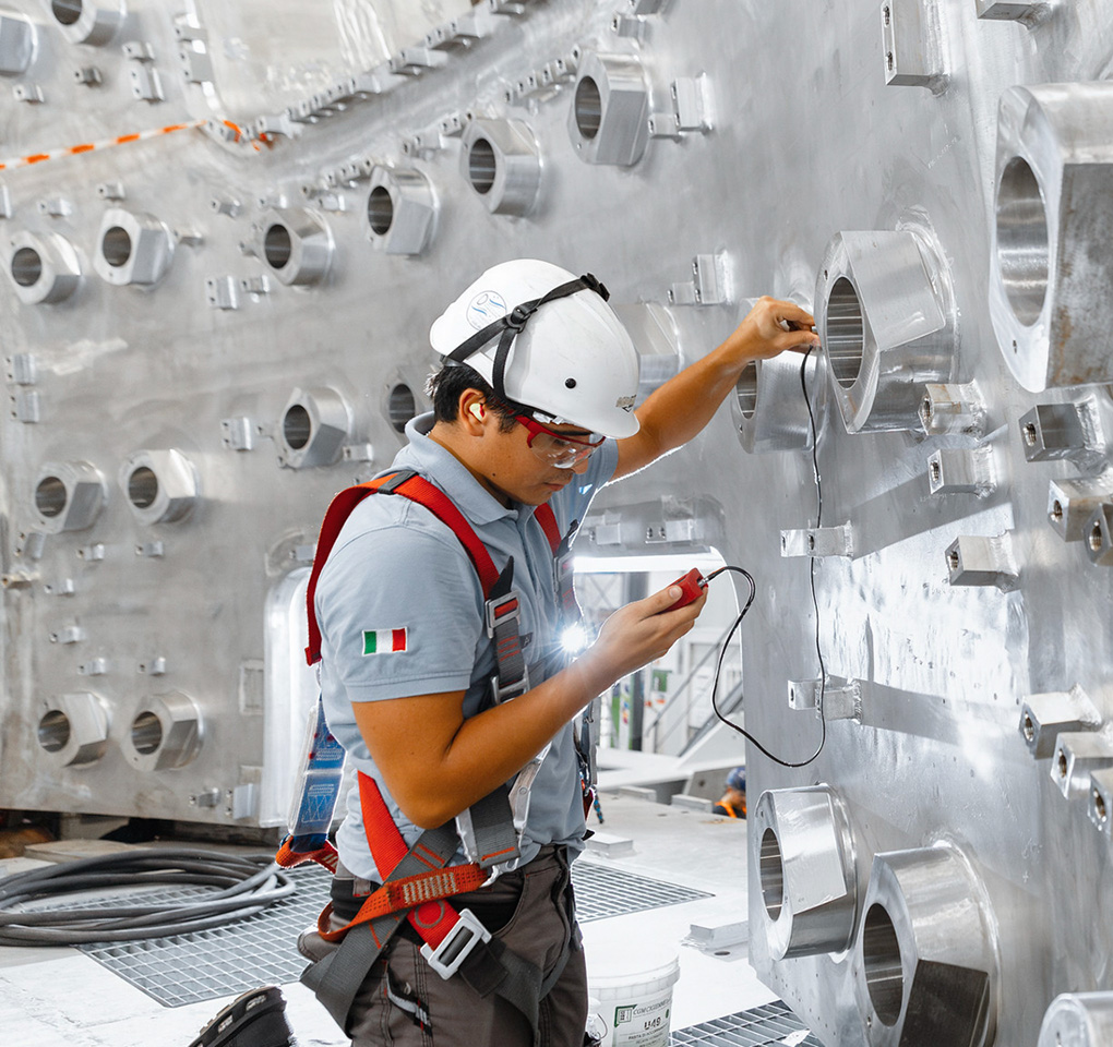 Un trabajador de la construcción con un casco rígido revisa un dispositivo de medición en su mano, con un fondo de una pared de metal con aperturas