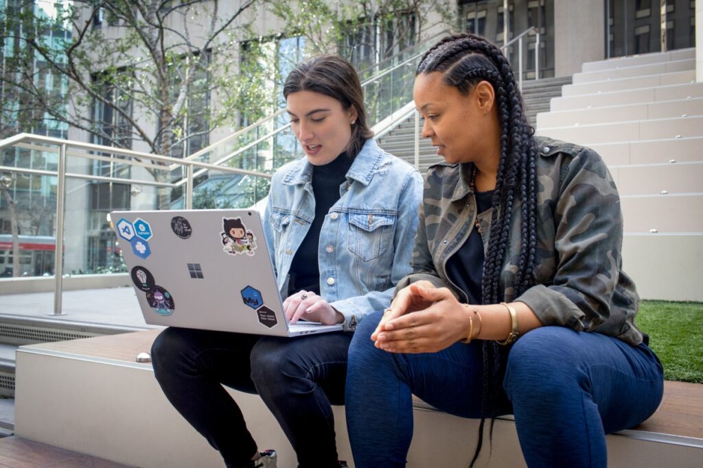 Dos personas sentadas en unos escalones de un edificio de oficinas trabajan juntas en una laptop