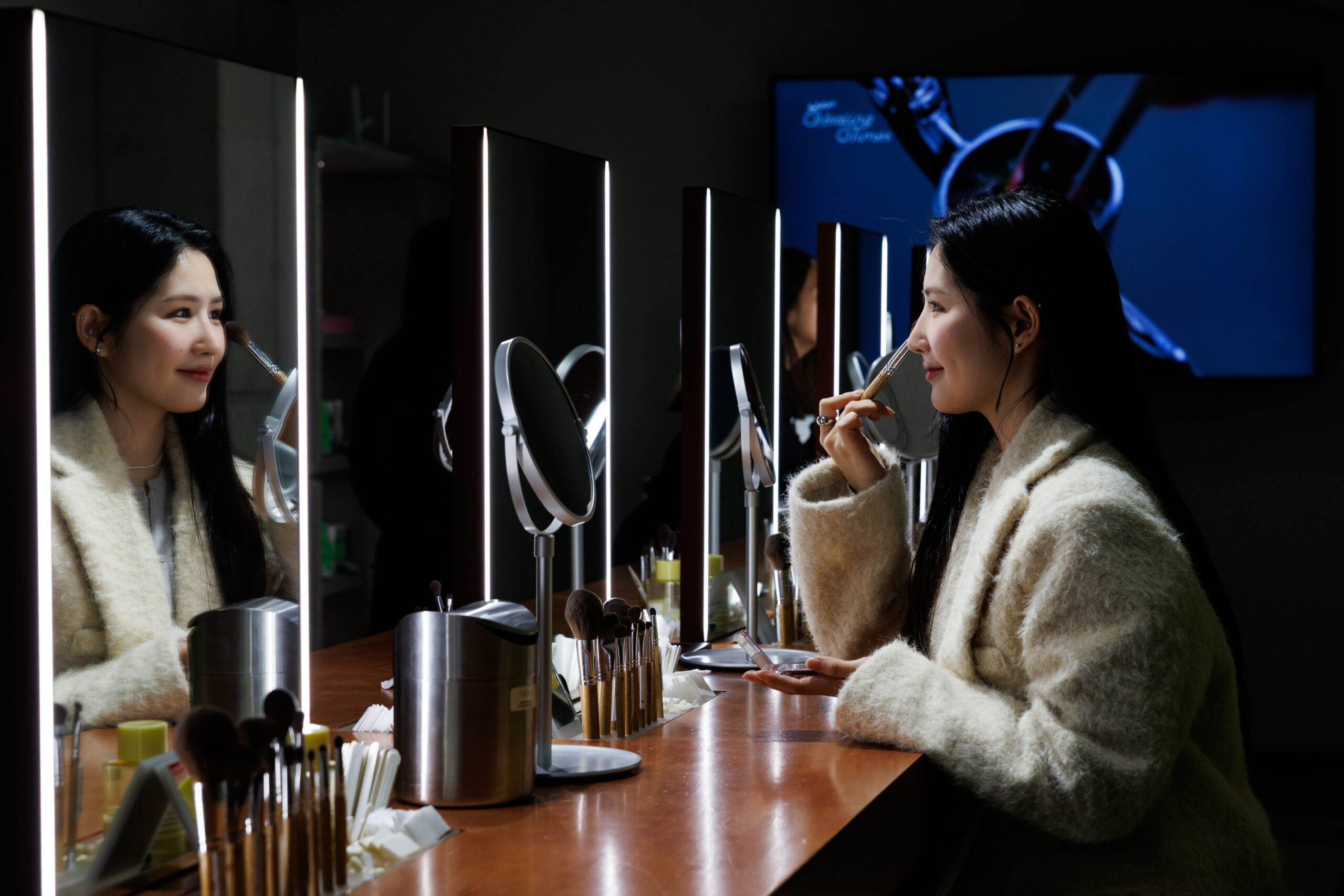 Una mujer con cabello negro largo con una chaqueta blanca se mira en el espejo mientras usa una brocha de maquillaje en su cara