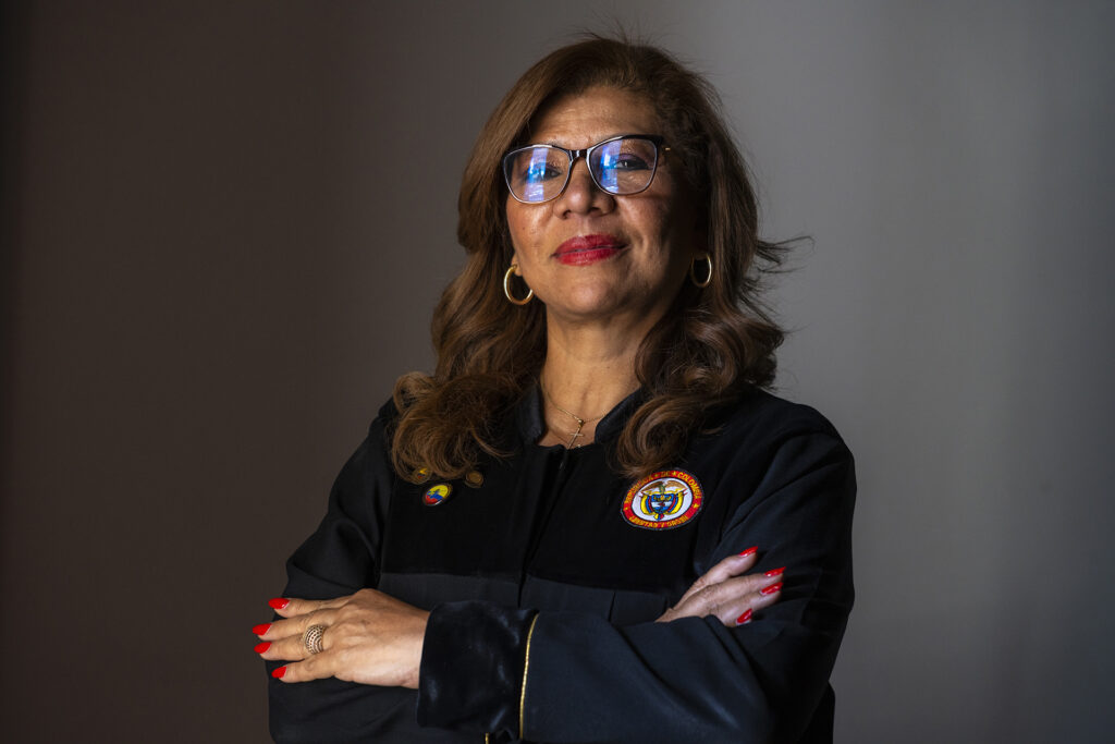 Uma mulher com toga de juíza e braços cruzados contra um fundo marrom escuro e cinza
