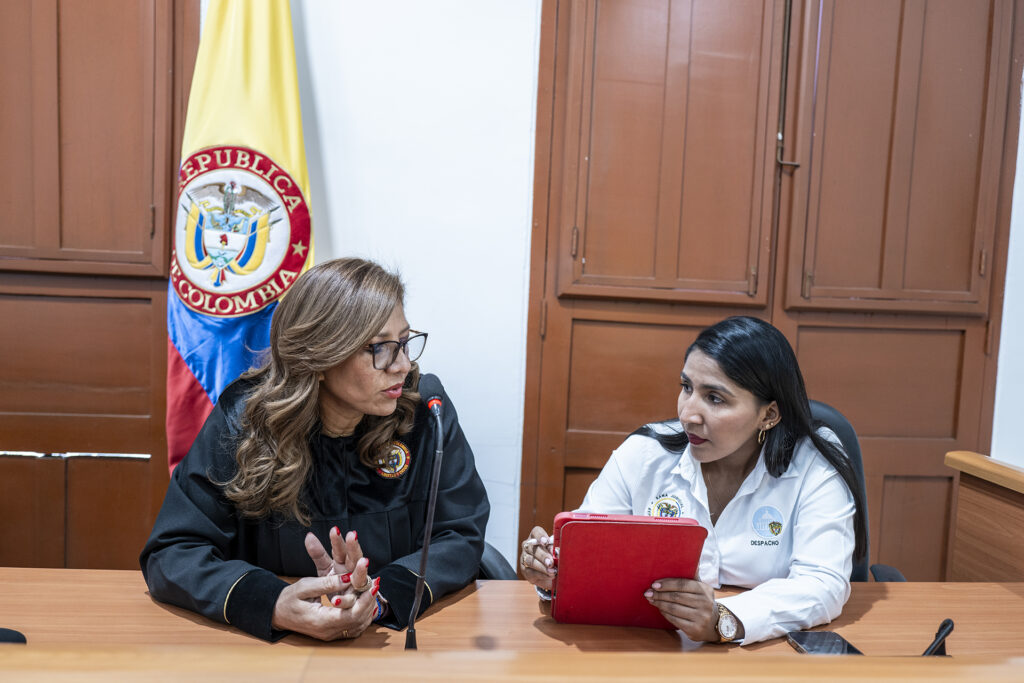 Uma mulher com vestes de juíza sentada em uma mesa com uma mulher segurando um tablet, com a bandeira colombiana ao fundo