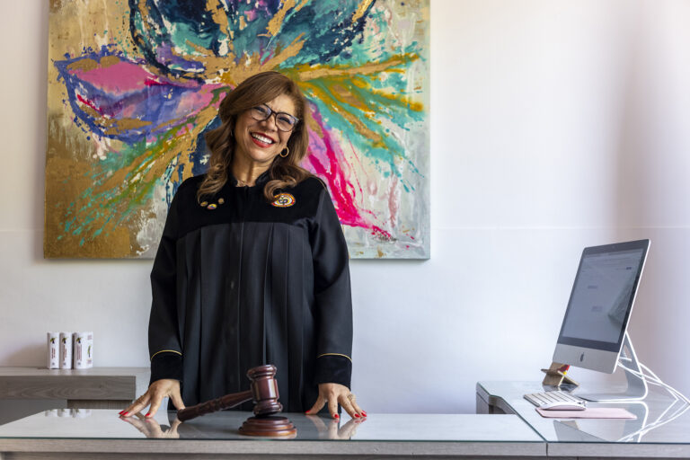 Uma juíza de toga está em pé diante de uma mesa com um martelo na frente dela, atrás dela uma pintura colorida e ao lado dela um computador