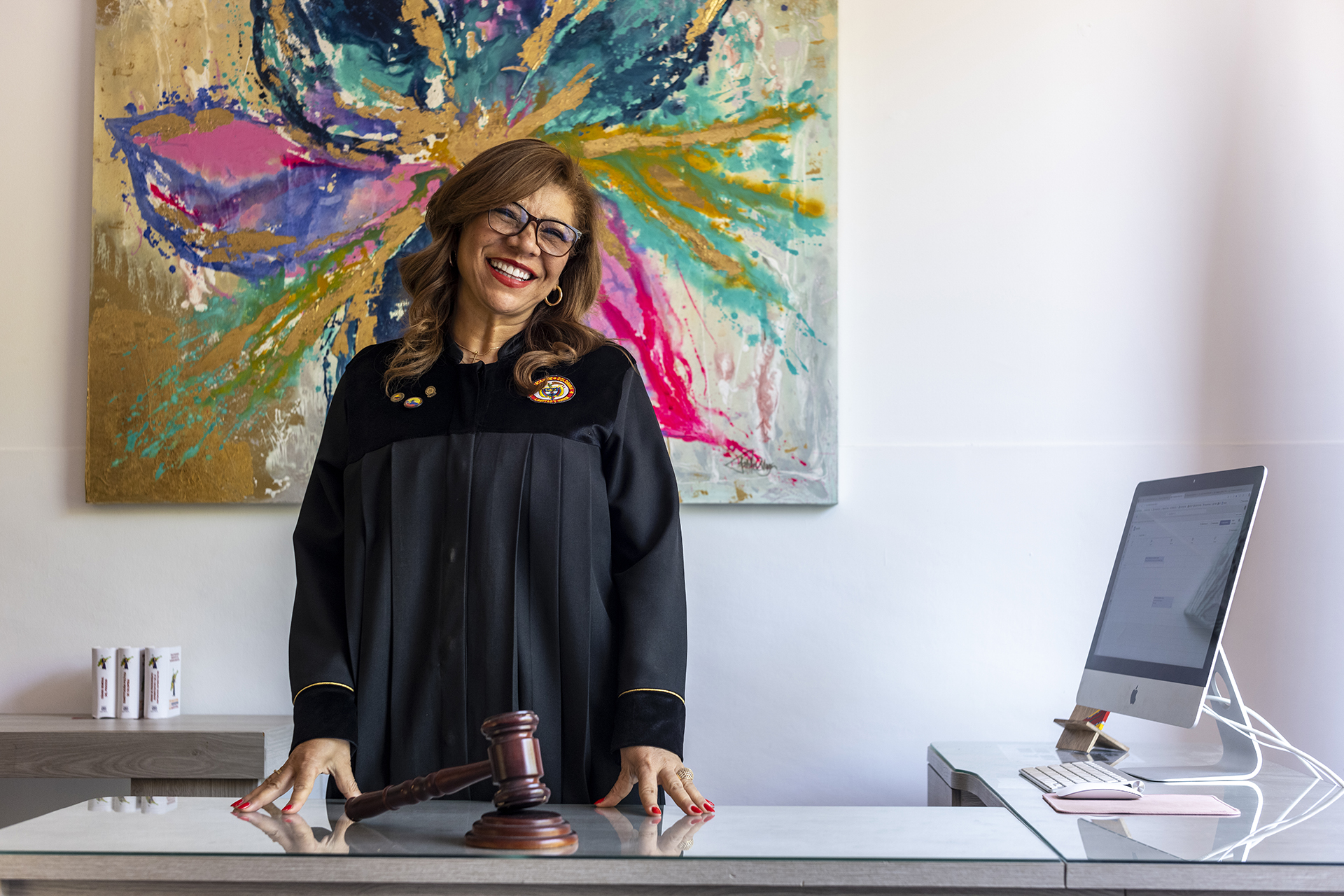 Uma juíza de toga está em pé diante de uma mesa com um martelo na frente dela, atrás dela uma pintura colorida e ao lado dela um computador