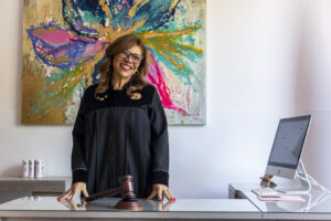 Una jueza con ropas judiciales está de pie en un escritorio con un mazo frente a ella, detrás de ella hay una pintura colorida y a su lado una computadora.