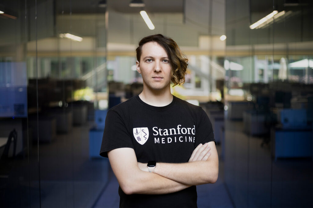 Un hombre con corte de cabello asimétrico, playera negra con las palabras "Stanford Medical", cruzado de brazos frente a unas puertas de cristal