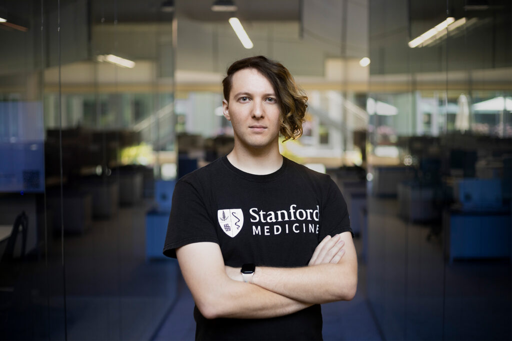 Un hombre con corte de cabello asimétrico, playera negra con las palabras "Stanford Medical", cruzado de brazos frente a unas puertas de cristal