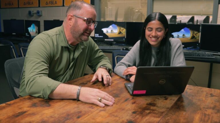 Un educador y una estudiante sentados en una mesa en un aula mientras observan la pantalla de una laptop