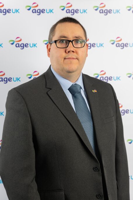 Un hombre con traje y corbata posa frente a un muro con el logo de Age UK