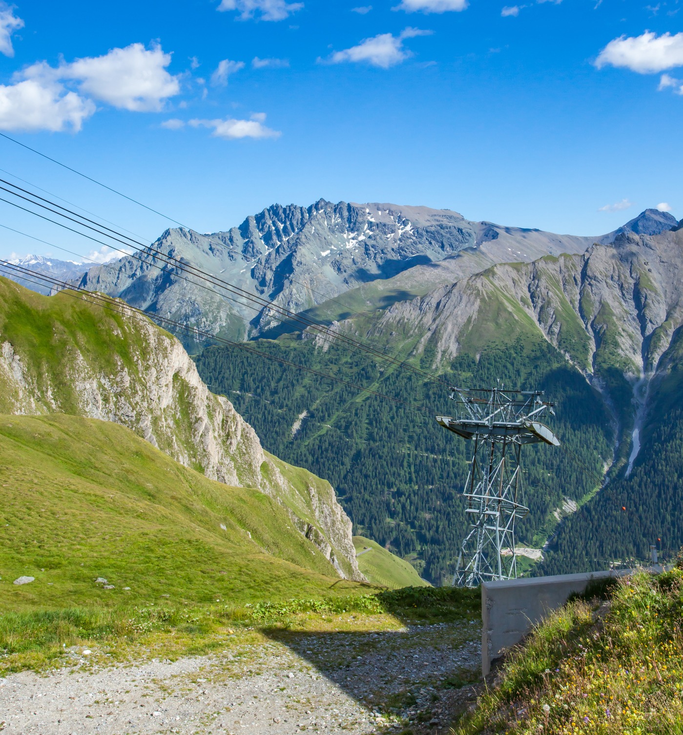 Una torre de teleférico y cables aéreos atraviesan un dramático valle alpino con montañas nevadas, praderas verdes y laderas boscosas bajo un cielo azul con nubes blancas.
